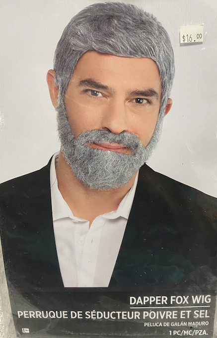 A gray faux-fur fox wig, shown on a man  with a faux-fur beard (not included) wearing a black blazer and white shirt. The product packaging text at the bottom reads 'DAPPER FOX WIG' and 'PERRUQUE DE SÉDUCTEUR POÍVRE ET SEL'. 1PC